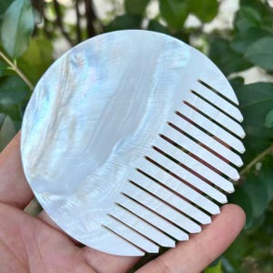 May include: A round, iridescent white comb with a pearlescent sheen. The comb features a series of parallel teeth on one side, and a smooth, rounded surface on the other. The comb is made of a natural material.