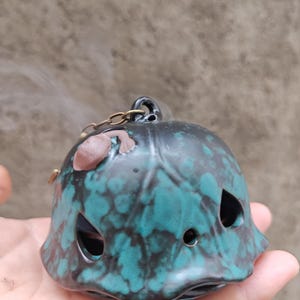 May include: A bell-shaped ceramic incense burner with a glossy black and teal mottled glaze. The object features small triangular cutouts, a small brown sculpted frog figure on the top, and a metal chain loop attached to the apex.