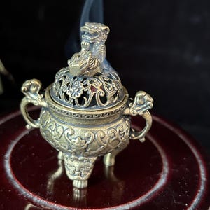 May include: A detailed brass incense burner with a decorative lid featuring a mythical creature. The burner has ornate handles and sits on three small legs. Wisps of smoke rise from the top, suggesting the burner is in use.