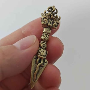 May include: A small, antique-style bronze dagger pendant. The pendant features a detailed handle with a multi-faced deity design and a lotus flower motif. The blade is triangular with a curved design.