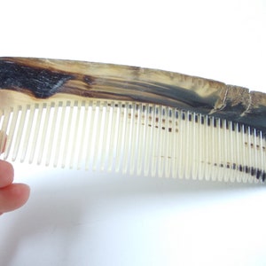 May include: A handcrafted comb with a natural horn handle and off-white teeth. The handle features a marbled pattern of dark brown and cream tones. The comb's teeth are closely spaced, and the overall design is simple and functional.