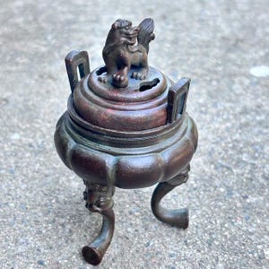 May include: Bronze incense burner with a detailed lion figure on the lid. The burner has a bulbous body, three curved legs, and two rectangular handles. The overall color is a dark, aged bronze.