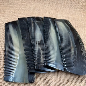 May include: A collection of black and gray combs with fine teeth. The combs have a marbled appearance and are arranged in a slightly overlapping pattern. The background is a textured, tan fabric.