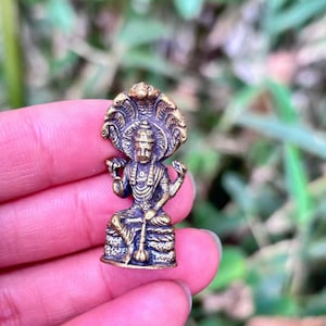 May include: A small, detailed brass statue of a seated deity, adorned with a multi-headed serpent halo. The figure is seated on an ornate base, holding objects in its hands. The statue has a weathered, antique appearance.