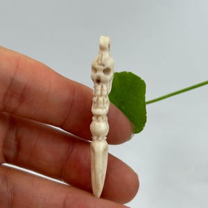 May include: A carved bone dagger pendant with a skull-like design at the top. The dagger is off-white and features intricate details along the handle. The pendant is held against a white background with a green leaf.