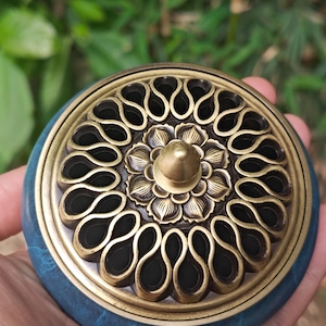 Puede incluir: Un quemador de incienso azul y latón con un diseño floral decorativo y una celosía intrincada. El quemador tiene una base redonda y una tapa con un pomo en la parte superior.
