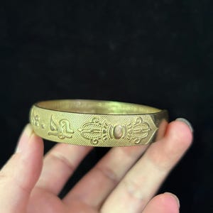 May include: A gold-colored bangle bracelet with etched patterns and symbols. The bracelet has a textured surface and is held in a hand against a dark background. The design includes repeating geometric shapes and what appears to be stylized text.