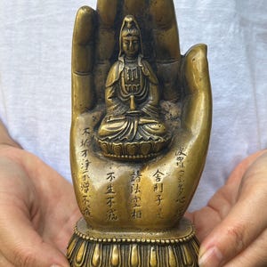May include: A bronze-colored statue of a seated figure within a hand-shaped base. The statue features intricate details, including the figure's serene expression and ornate clothing. The hand-shaped base has inscriptions.