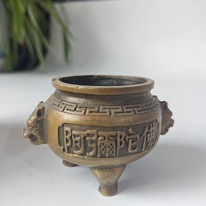 May include: A bronze-colored incense burner with three legs and a circular opening. The burner features embossed Chinese characters and lion head handles. The design includes a decorative band around the rim.