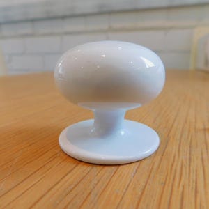 May include: A white, oval cabinet knob with a rounded base and a matching screw. The knob is glossy and sits on a wooden surface. Suitable for drawers and cabinets. The knob is approximately 4 cm in diameter.