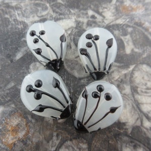 May include: Four white glass beads with black floral designs. The beads are teardrop shaped and have a glossy finish.