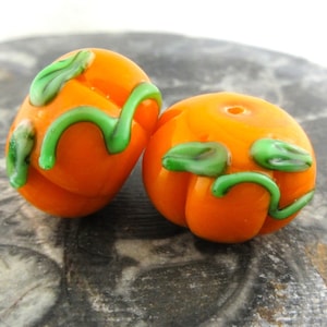 May include: Two orange glass pumpkin beads with green leaves and vines. The beads are about 1 inch in diameter.