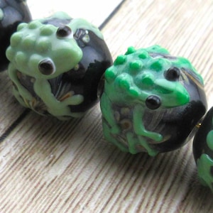 May include: Close-up of green frog beads with black and floral accents. The frogs have detailed features and dark eyes. The beads are arranged on a light-colored wooden surface.