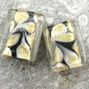 May include: Two rectangular glass beads with a white, black, and gold marbled pattern. The beads are translucent and have a glossy finish.