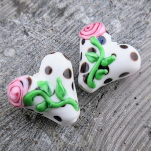 May include: Two white heart-shaped glass beads with black polka dots and green and pink floral accents.
