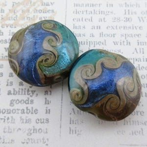 May include: Two round, dichroic glass beads with swirling, abstract designs. The beads feature a blend of blue, teal, and brown hues with a metallic sheen. The swirling patterns are in a light beige color. These beads are ideal for jewelry making.
