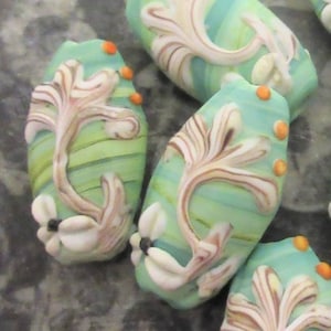May include: A set of five green, white, and brown glass beads with a swirling pattern and white floral accents. The beads are oval shaped and have a glossy finish.