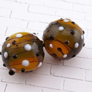 May include: Two round glass beads with a mix of colors. The beads are primarily amber and olive green, with white and black polka dots. The beads have a glossy finish and are set against a white brick background.