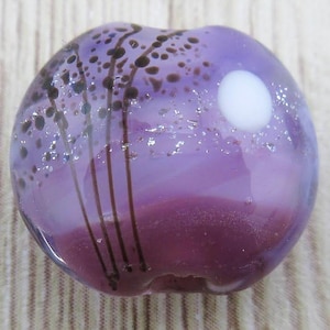 May include: Purple glass bead with a white moon and brown lines, resembling a night sky with trees and a moon.