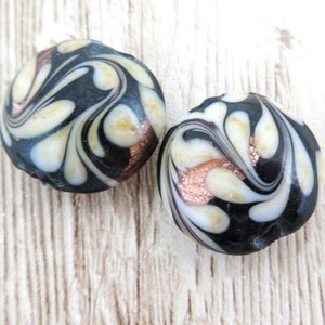 May include: Two round glass beads with a black, white, and copper swirl pattern. The beads are approximately 1 inch in diameter.