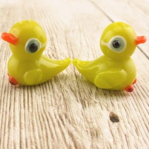 May include: Two small yellow glass bird figurines with red beaks and blue eyes. The birds are sitting on a wooden surface.