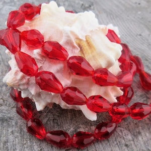 May include: A strand of faceted, teardrop-shaped red beads is draped over a white seashell. The beads are strung together, creating a necklace-like arrangement. The beads have a glossy finish and reflect light, adding a touch of elegance.