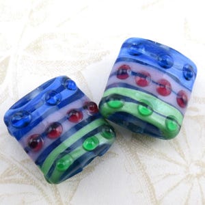 May include: Two square glass beads with a striped design in blue, green, red, and lavender. Each bead has several raised, round, colored accents. The beads are on a white patterned surface.