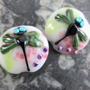 May include: Two white glass beads with a green and black dragonfly design. The dragonflies have turquoise accents and are surrounded by pink and purple flowers.