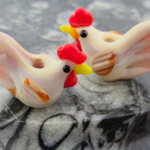 May include: Two white ceramic chicken figurines with red combs and yellow beaks. The chickens have brown and white striped wings and a hole in the back for stringing.
