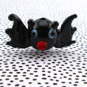 May include: A black glass bat figurine with blue eyes and a red nose. The bat has outstretched wings and small ears. The figurine is set against a white background with black polka dots.