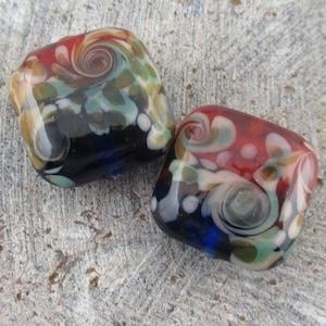 May include: Two square glass beads with a colorful swirl pattern. The beads are red, blue, green, white, and black.