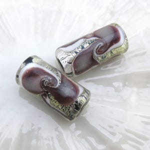 May include: Two handmade glass beads with a swirling design. The beads feature a mix of colors, including burgundy, white, silver, and gold. The cylindrical beads are displayed on a white surface.