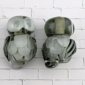 May include: Two glass owl figurines featuring a translucent grey and white color palette. The left owl displays a white face and chest with grey wings, while the right owl shows a darker grey head and body with white feather-like patterns.