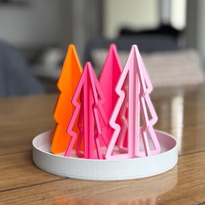 Könnte beinhalten: Eine dekorative Anordnung von farbenfrohen, stilisierten Weihnachtsbäumen. Die Bäume sind in Orange-, Rosa- und Hellrosa-Tönen gehalten und stehen auf einem weißen, runden Tablett. Die Bäume haben ein modernes, geometrisches Design für die Feiertagsdekoration.