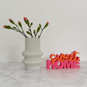 Op de afbeelding: Een grijze, geribbelde vaas met roze en groene bloemen staat naast een roze en oranje "Sweet Home" bord. De vaas heeft een uniek ontwerp en het bord voegt een decoratief tintje toe.