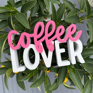 May include: A white and pink "Coffee Lover" sign. The word "coffee" is in a cursive pink font, and the word "lover" is in a block white font. The sign is set against a backdrop of green leaves and small yellow fruits.