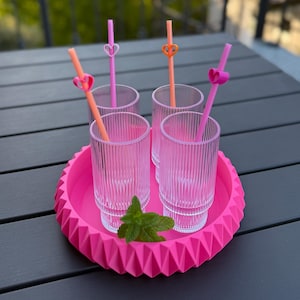 Könnte beinhalten: Vier klare, geriffelte Trinkgläser mit rosa Strohhalmen und herzförmigen Verzierungen, angeordnet auf einem hellrosa Tablett mit geometrischen Kanten. Ein Minzzweig sorgt für einen frischen Akzent. Bereit für ein erfrischendes Getränk.