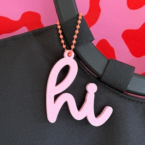 Könnte beinhalten: Ein hellrosa Acryl-Anhänger in Form des Wortes hi in Schreibschrift. Der Anhänger ist mit einer kleinen orangefarbenen Kugelkette an einem schwarzen Taschengriff befestigt. Die Oberfläche des Anhängers hat eine matte Textur und eine hellrosa Farbe.