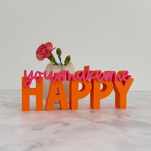 Könnte beinhalten: Ein dekoratives Schild in Orange und Pink mit der Aufschrift "you make me HAPPY". Vor dem Schild steht eine kleine weiße Vase mit einer rosa Blume und grünen Knospen. Die Buchstaben sind 3D und stehen aufrecht.