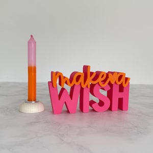 Könnte beinhalten: Eine rosa-orangefarbene Ombre-Kerze in einem weißen Halter neben einem rosa-orangefarbenen Holzschild mit der Aufschrift "make a wish". Die Kerze ist angezündet und das Schild ist in einer verspielten Schriftart.
