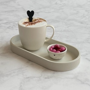 Op de afbeelding: Een beige keramische mok met verticale ribbels, gevuld met cappuccino en een zwarte hartvormige roerder, staat op een bijpassend ovaal dienblad. Een kleine kom met roze snoepjes staat ook op de dienblad. Marmeren achtergrond.
