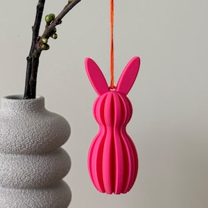 May include: A bright pink, ribbed bunny ornament hanging from an orange string. The bunny is suspended in front of a neutral background, with a vase and a branch with buds visible on the left. The ornament is a decorative item.