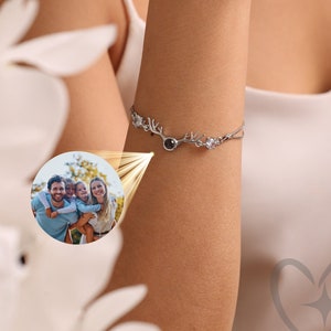 Op de afbeelding: Een zilveren armband met een hertengeweimotief en een kleine, ronde zwarte steen. De armband wordt om een arm gedragen, met een familiefoto in een cirkelvormige inzet. De achtergrond is wazig, met witte bloemen op de voorgrond.