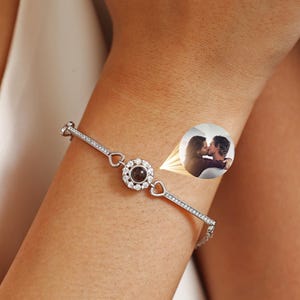 Könnte beinhalten: Ein silbernes Armband mit einem zentralen runden Anhänger und einer Reihe kleiner, klarer Steine. Der Anhänger scheint ein kleines Bild eines Paares zu enthalten. Das Armband befindet sich am Handgelenk einer Person, mit weißem Stoff im Hintergrund. Das Logo "myLoveSparks" ist sichtbar.