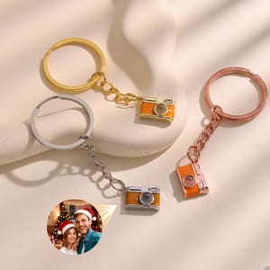 May include: Three keyrings with miniature orange camera charms. One keyring is gold, one is silver, and one is rose gold. Each camera charm has a silver lens and a chain connecting it to the keyring. A small photo is attached to the silver keyring.