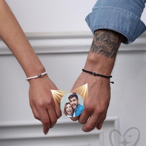 May include: Two people holding hands with a photo of a couple inside a circle on their wrists. The photo is surrounded by a golden glow. The background is a white wall.