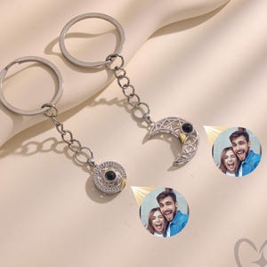 May include: Two silver keychains with a circular and crescent moon pendant. The pendants feature a photo of a couple. The keychains are connected to a chain and a keyring. The background is a light beige color.