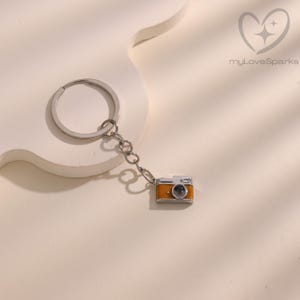 May include: A silver keychain with a small, orange and silver camera charm. The camera charm has a black lens and is attached to the keychain with a small chain. The background is a light beige color.