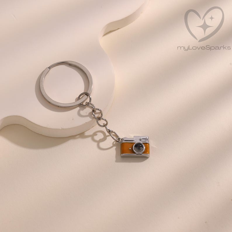 May include: A silver keychain with a small, orange and silver camera charm. The camera charm has a black lens and is attached to the keychain with a small chain. The background is a light beige color.