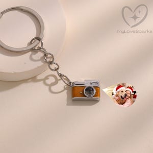 Peut inclure: Un porte-clés avec un charm en forme de mini appareil photo orange et argenté. Le charm est attaché à une chaîne et à un anneau. Une petite photo est projetée depuis l'appareil photo. Le logo "myLoveSparks" est en haut à droite.